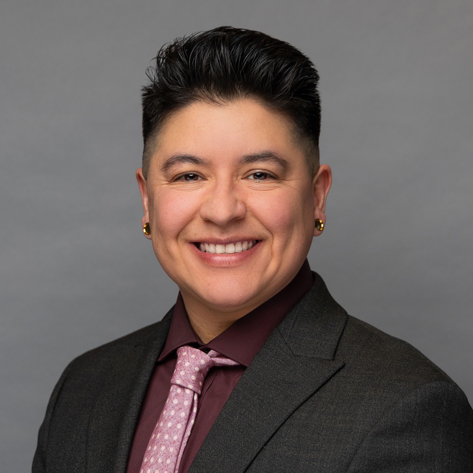 A headshot of Dallas Aguilera Martinez with a warm smile, wearing a dark grey blazer, burgundy shirt, and a pink patterned tie. They have short, styled hair, and wear gold, tunnel plug earrings
